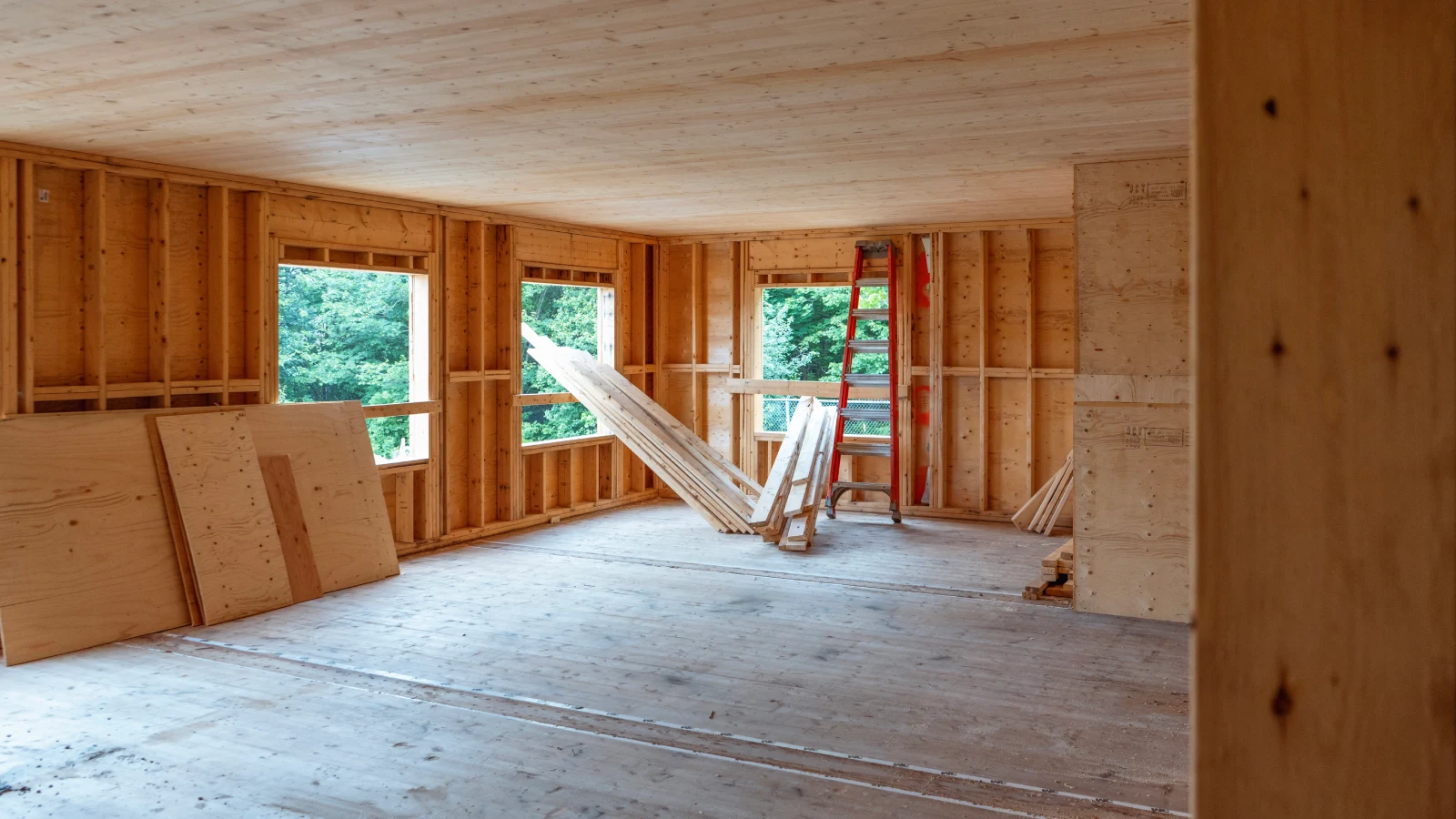 An unfinished interior of a building under construction. The room features exposed wooden framework and plywood walls, with three large window openings letting in natural light.