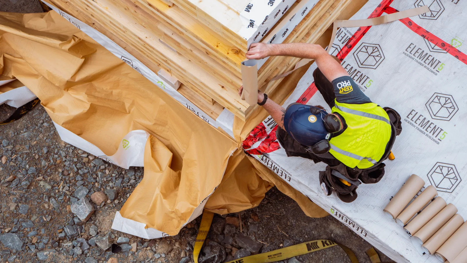 An overhead view of a construction worker examining a stack of lumber. They are wearing a hard hat and hear protection and a reflective vest.