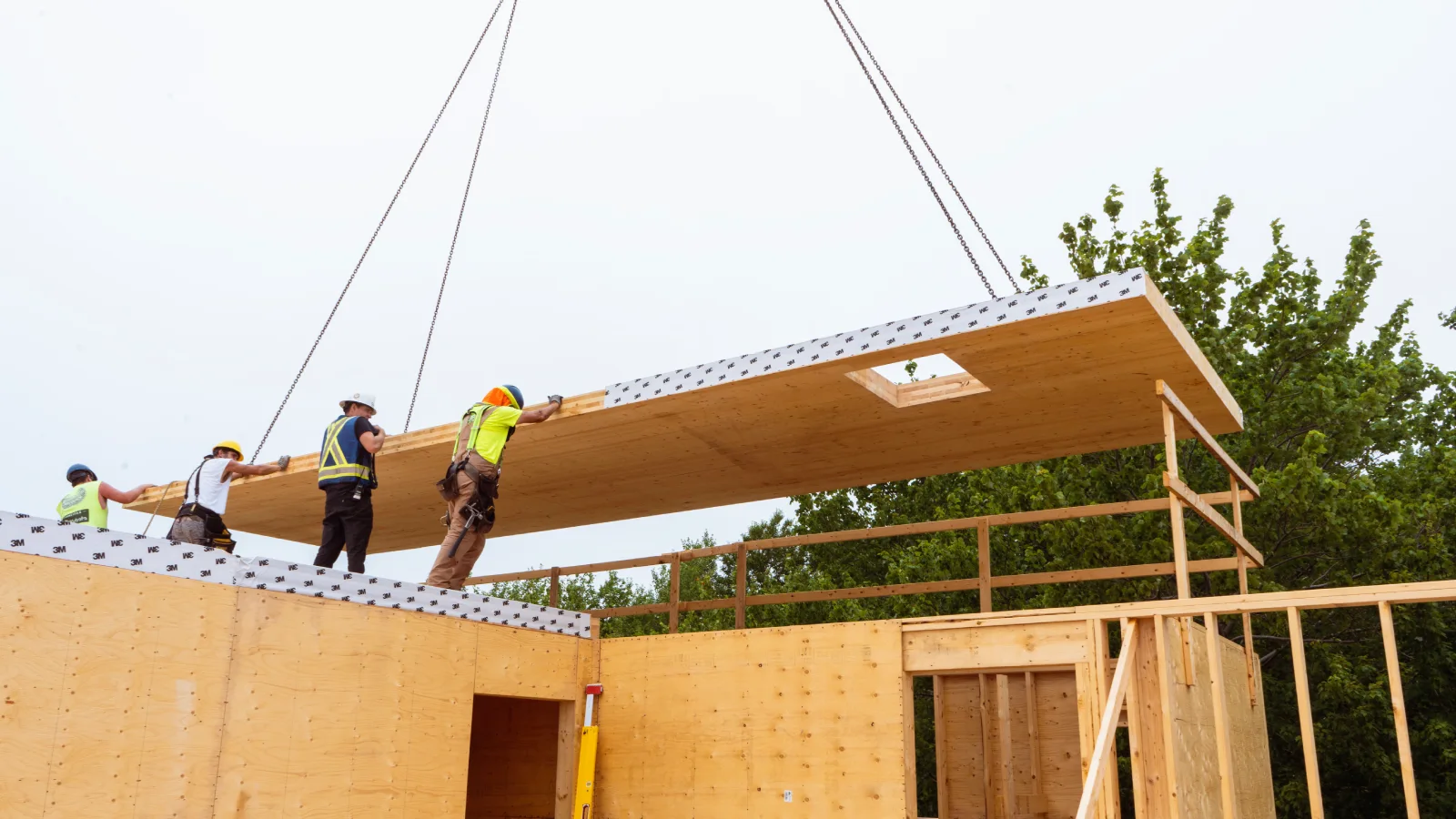 A crane is lowering a large section of wood into the framing of a wooden structure. 4 construction workers are guiding it into place. They are wearing hard hats and reflective clothing. In the background are the leaves of a large tree and grey blue sky.
