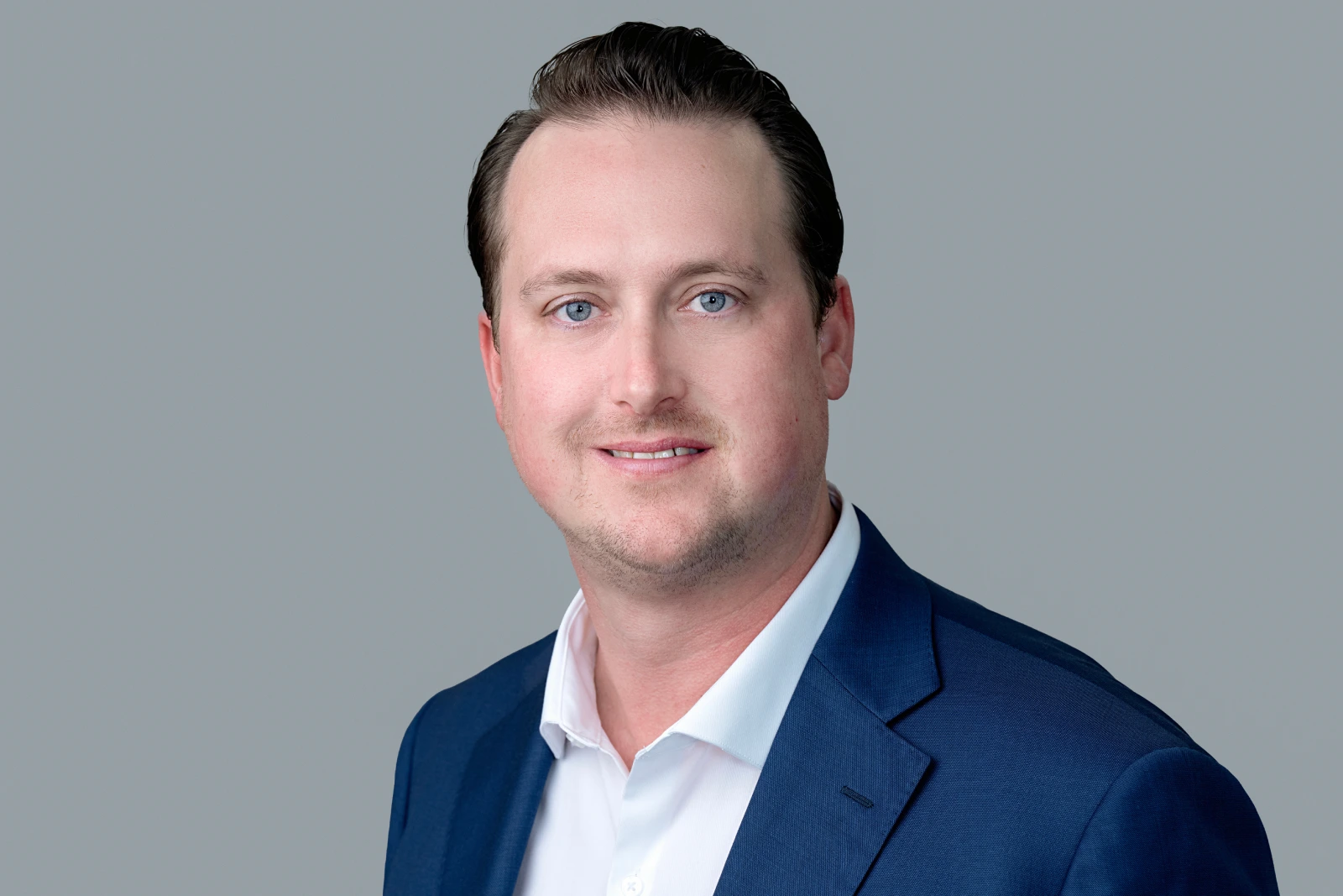 This is a portrait of a man wearing a blue suit jacket and white shirt. He is smiling and hair is neatly combed. The background of the image is a plain grey color.