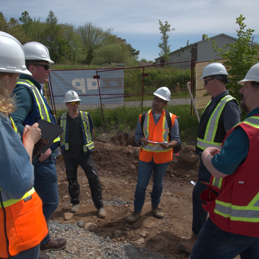 Group of construction workers and engineers wearing hard hats and high-visibility vests stand on a gravel site, engaged in a discussion. One person holds a clipboard while another takes notes on a tablet, with a fenced-off area and trees in the background.
