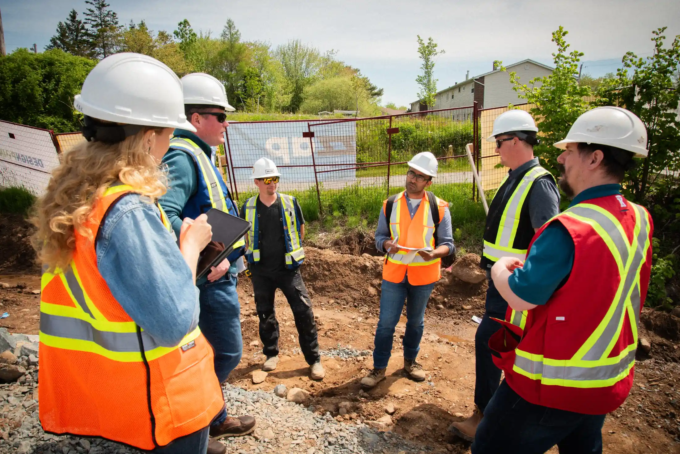 Group of construction workers and engineers wearing hard hats and high-visibility vests stand on a gravel site, engaged in a discussion. One person holds a clipboard while another takes notes on a tablet, with a fenced-off area and trees in the background.